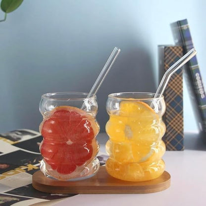 Two glass cups with fruit slices and straws on a wooden coaster.