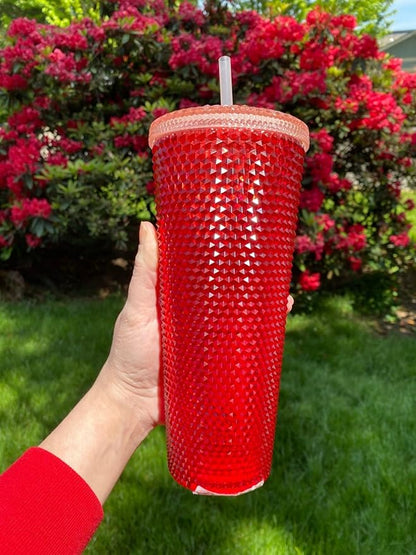 Red textured tumbler with a beige lid held in front of a flowering bush.