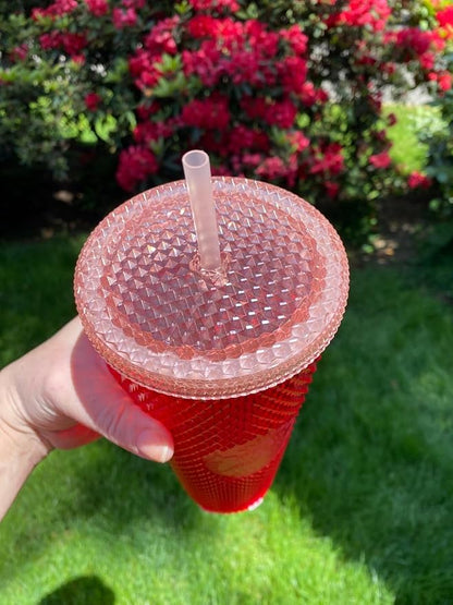 Red cup with a textured lid held in front of green grass and pink flowers